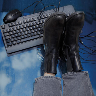 Black boots are lying on a table with a keyboard and a computer mouse. Many cables are visible. | © Valentina Schuster