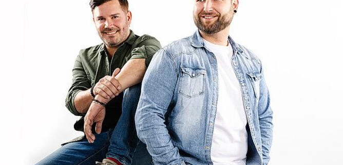 Two men are happily smiling in front of a white background. They are wearing casual clothing and standing in a relaxed pose. | © FG Event Productions OG