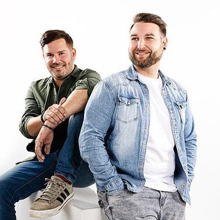 Two men are happily smiling in front of a white background. They are wearing casual clothing and standing in a relaxed pose. | © FG Event Productions OG