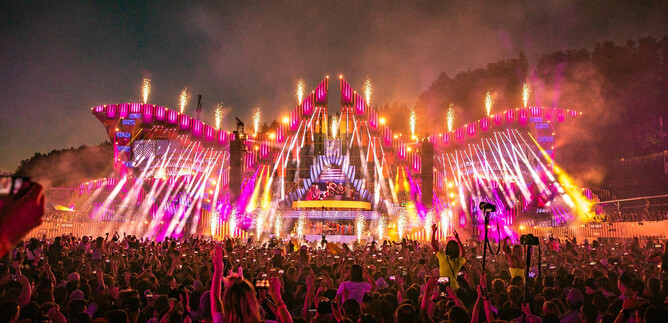 A lively music festival with a huge stage surrounded by colorful lights and fireworks. The crowd cheers and is full of energy. | © Geoffrey Hubbel