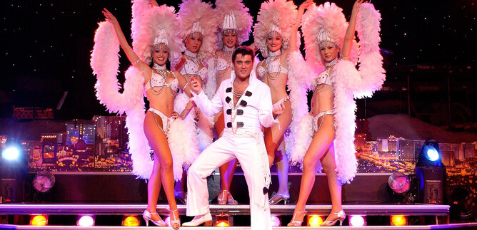 A stage performance with dancers in glamorous costumes and feather boas. In the center stands a man in a white suit, surrounded by six female dancers. | © COFO Entertainment GmbH & Co.KG