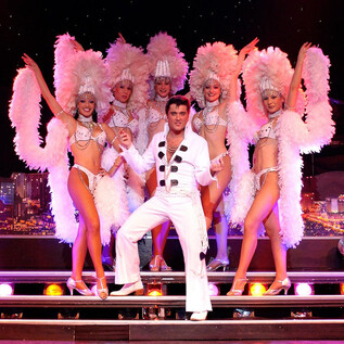 A stage performance with dancers in glamorous costumes and feather boas. In the center stands a man in a white suit, surrounded by six female dancers. | © COFO Entertainment GmbH & Co.KG