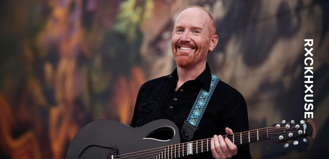 A smiling man is holding a guitar in his hand. In the background, colorful, abstract patterns can be seen. | © Lisa Sciliano