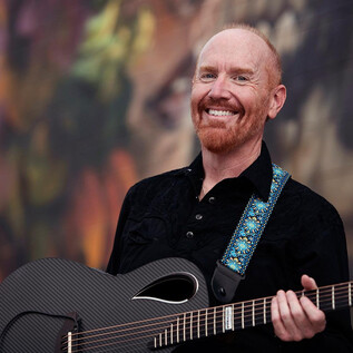 A smiling man is holding a guitar in his hand. In the background, colorful, abstract patterns can be seen. | © Lisa Sciliano