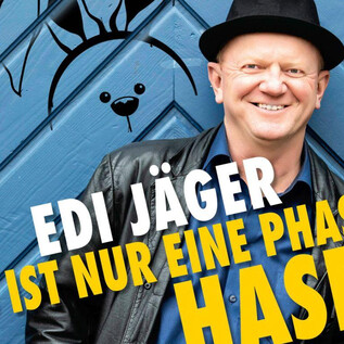 A smiling man is wearing a black hat and a black jacket in front of a blue wall. In the background, cartoon-like rabbit illustrations can be seen. | © Kurt-Sixtl_Neuhofen
