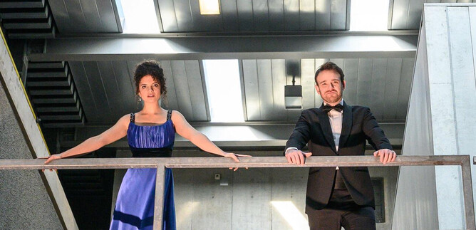 An elegant couple stands on a balcony in a modern building. They are dressed in formal evening wear and are looking forward. | © SLT / Tobias Witzgall