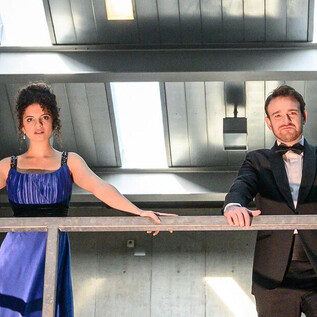 An elegant couple stands on a balcony in a modern building. They are dressed in formal evening wear and are looking forward. | © SLT / Tobias Witzgall