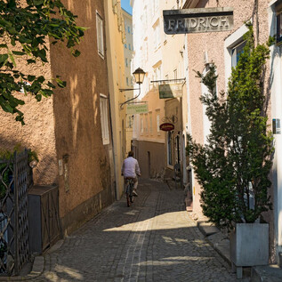 A narrow alley with colorful houses and a cyclist. Trees and a sign with the name "Friedrich" are also visible. | © TSG Tourismus Salzburg GmbH