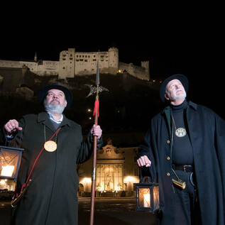 Two men in historic garments stand at night with lanterns in front of a illuminated castle. They carry a walking stick and a large axe and look seriously into the distance. | © Reischenböck & Knoll