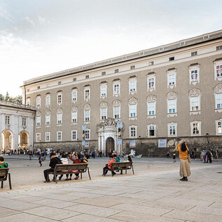 A group of men stands together, expressive and serious. They hold spears and look into a dark, cloudy environment. | © DomQuartier Salzburg
