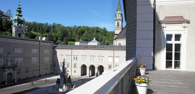 Una hermosa vista de una plaza histórica con edificios y una fuente. En el fondo hay colinas verdes y el cielo está claro y azul. | © DomQuartier