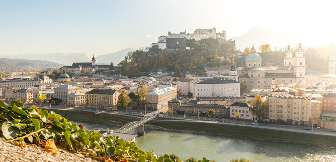 Una vista pintoresca de la ciudad de Salzburgo con la fortaleza de Hohensalzburg en el fondo. El río Salzach atraviesa la ciudad, rodeado de suaves colinas y luz dorada. | © AdobeStock