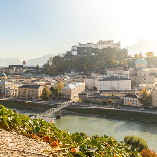 A picturesque city view of Salzburg with the Hohensalzburg Fortress in the background. The Salzach River flows through the city, surrounded by gentle hills and golden light. | © AdobeStock