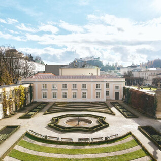 A beautiful garden with symmetrical hedges and a central fountain. In the background, buildings and a clear, blue sky are visible. | © Salzburg Museum/Tolga Salpat