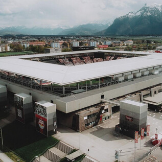 A modern football stadium with a spacious grandstand. In the background, mountains and an urban landscape can be seen.