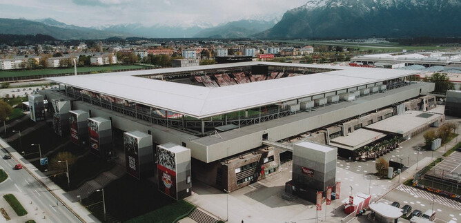 A modern stadium with a large roof and seating. In the background, mountains and a cityscape can be seen.
