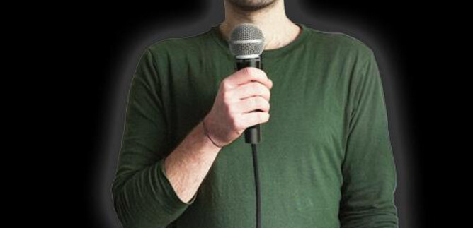 A man with a microphone stands in front of a black background. He is wearing a green shirt and looking at the camera. | © Fabi Rommel