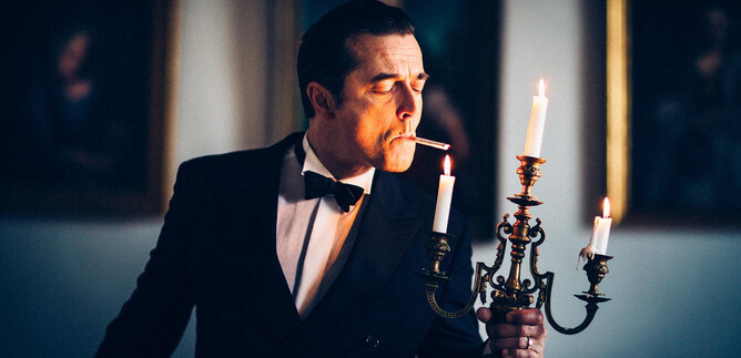 An elegantly dressed man in a tuxedo holds a candelabrum with burning candles. He is smoking a cigarette and stands in a room with classical paintings in the background. | © Marcel Klette