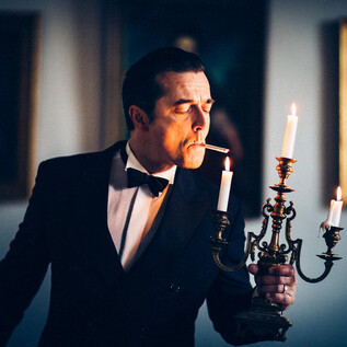 An elegantly dressed man in a tuxedo holds a candelabrum with burning candles. He is smoking a cigarette and stands in a room with classical paintings in the background. | © Marcel Klette