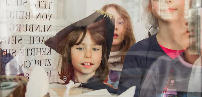 A group of children curiously looks through a window. They are in festive clothing and are observing objects displayed on a table. | © Eva trifft