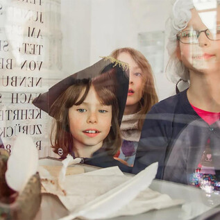 A group of children curiously looks through a window. They are in festive clothing and are observing objects displayed on a table. | © Eva trifft