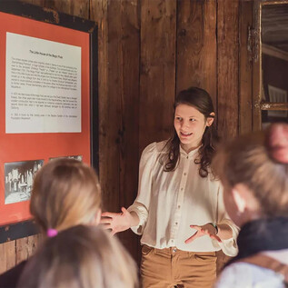 A young woman is explaining something to a group of listeners in front of a whiteboard. Historical images and texts are visible in the background. | © Eva trifft