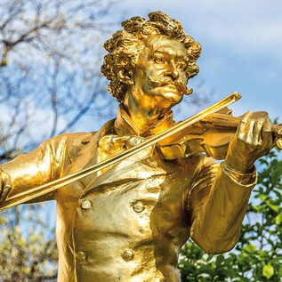 A golden statue of a violinist happily plays his instrument. The background features green trees and a clear sky. | © AlexejFedorenko/Shutterstock, Moises Patrici