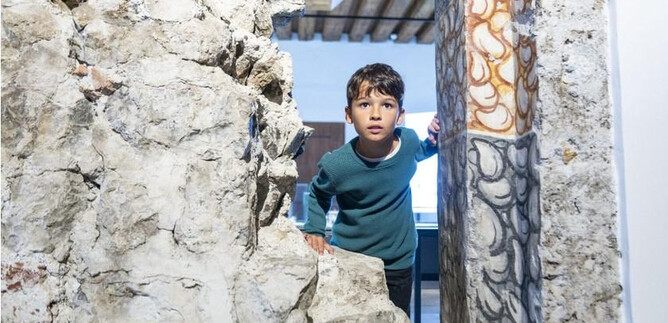 Un niño mira con curiosidad a través de una abertura en la pared. En el fondo se puede ver una habitación iluminada. | © Salzburg Museum