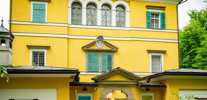 A pretty yellow building with green shutters. It has an inviting atmosphere and is surrounded by trees. | © Melanie Wressnigg