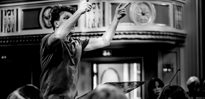 A conductor leads the orchestra with energetic gestures. The scene is in black and white, which emphasizes the drama of the performance. | © Salzburger Kulturvereinigung