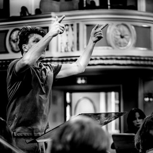 A conductor leads the orchestra with energetic gestures. The scene is in black and white, which emphasizes the drama of the performance. | © Salzburger Kulturvereinigung