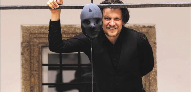 A man smiles and holds a black mask that is hanging on a stand. In the background, a building with windows can be seen. | © Erika Mayer