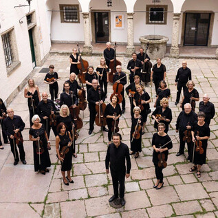 A large orchestra group is standing in the courtyard of a building. The musicians are wearing black clothing and holding their instruments. | © Salzburger Kulturvereinigung