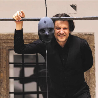 A man in black stands with a smile next to a hanging mask. In the background, an old wall and a window can be seen. | © Erika Mayer