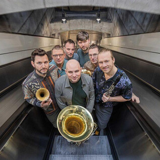A group of seven musicians stands in a modern environment. They hold various wind instruments and pose on an escalator. | © Severin Koller