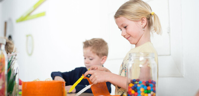 Two children are working creatively at a table. One of the children is cutting orange material, while the other is playing with colorful beads. | © Hannelore Kirchner