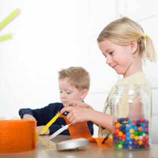 Deux enfants travaillent de manière créative à une table. L'un des enfants coupe du matériel orange, tandis que l'autre joue avec des perles colorées. | © Hannelore Kirchner