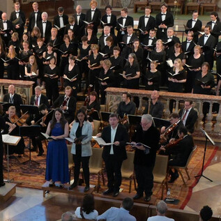 A choir is standing on a stage in a church and singing. Musicians with instruments are also visible on the stage. | © Dom zu Salzburg