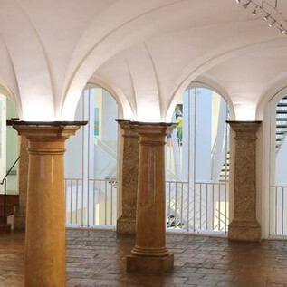 A bright exhibition space with columns and artworks on the walls. In the background, stairs and other design elements are visible. | © Stadtgalerie Rathaus