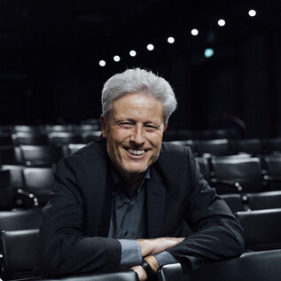 A smiling man in a suit sits in an empty theater. In the background, empty rows of seats are visible. | © Sky Sport Austria