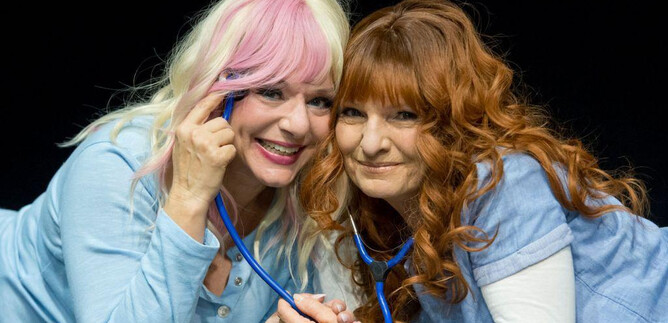 Two women in blue shirts are lying next to each other on stage. They are laughing and holding a stethoscope, indicating a medical or humorous performance. | © Christian Treweller