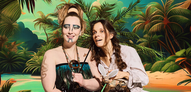 Two women are happily posing in front of a tropical background with palm trees. One woman is holding a coconut, while the other is showcasing animal accessories. | © Mathias Schuh