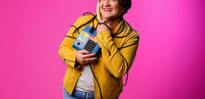 A cheerful woman is holding a blue telephone and smiling. The background is bright pink. | © Stefan Joham