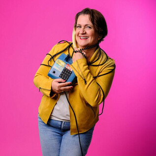 A cheerful woman is holding a blue telephone and smiling. The background is bright pink. | © Stefan Joham