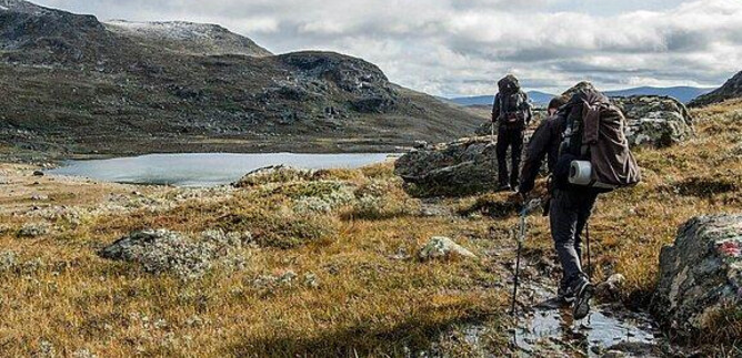 Two hikers are traversing a green, hilly landscape with some rocks and a calm lake in the background. The sky is cloudy, and the scenery appears wild and untouched. | © Messezentrum Salzburg