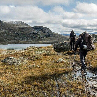 Two hikers are traversing a green, hilly landscape with some rocks and a calm lake in the background. The sky is cloudy, and the scenery appears wild and untouched. | © Messezentrum Salzburg