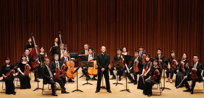 An orchestra is standing on a stage. The conductor is at the front, surrounded by musicians with various instruments. | © COLUMBUS Reisen GmbH & Co.KG