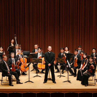 An orchestra is standing on a stage. The conductor is at the front, surrounded by musicians with various instruments. | © COLUMBUS Reisen GmbH & Co.KG