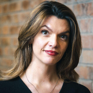 A woman with long, curly hair stands in front of a brick wall. She has a friendly expression and is wearing a black top. | © Ingo Pertramer