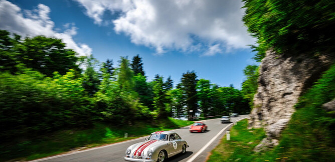 A classic car with racing stripes drives along a winding road. Surrounded by green trees and a blue sky with clouds. | © motorsportpics.at